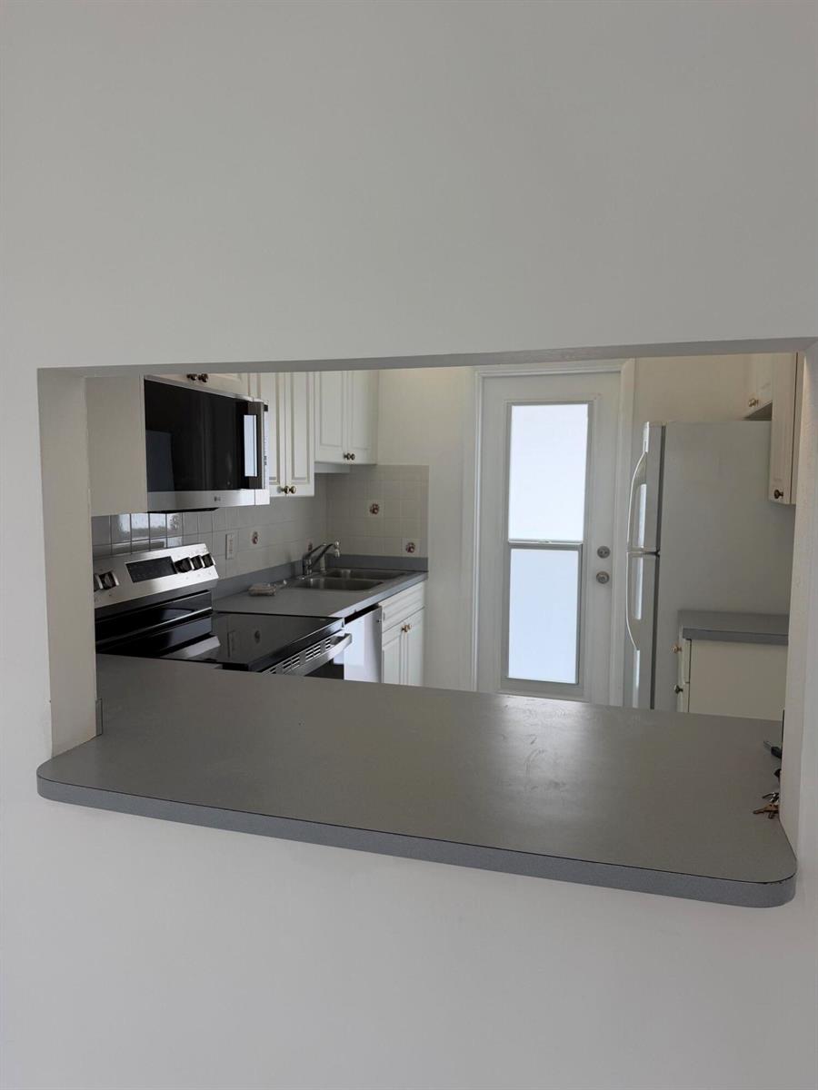 A kitchen viewed through a pass-through window features white cabinets, gray countertops, and stainless appliances — 1100 Pine Drive # 102, Pompano Beach FL 33060, POMPANO BEACH, FL