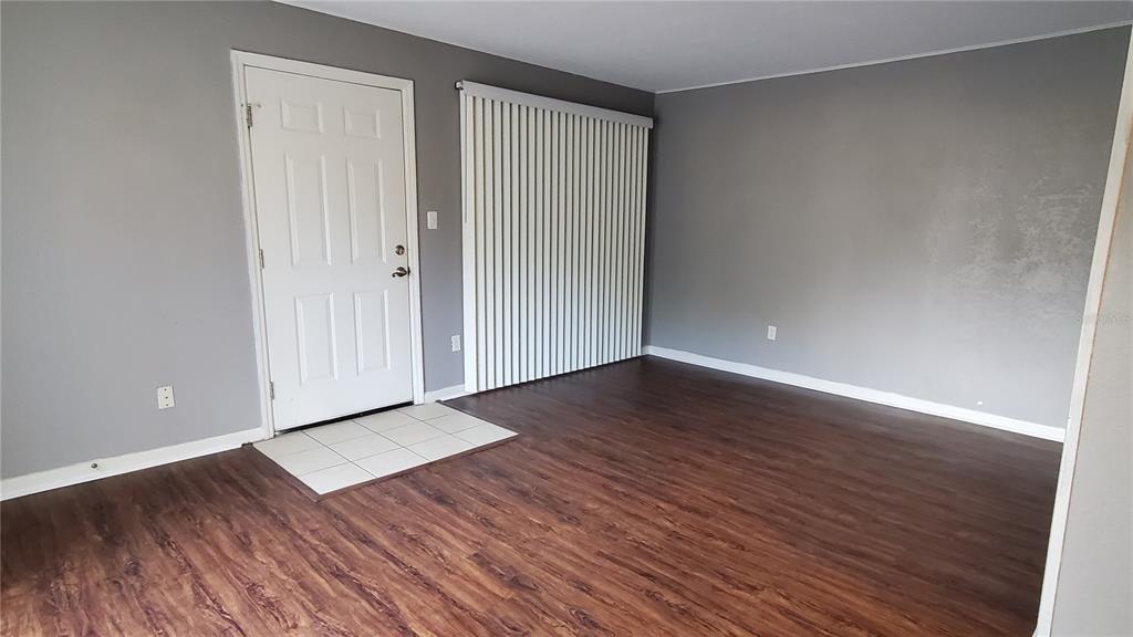 An empty room features gray walls, wood-look flooring, a white entry door, and vertical blinds — 507 NW 39TH RD UNIT 237, GAINESVILLE, FL