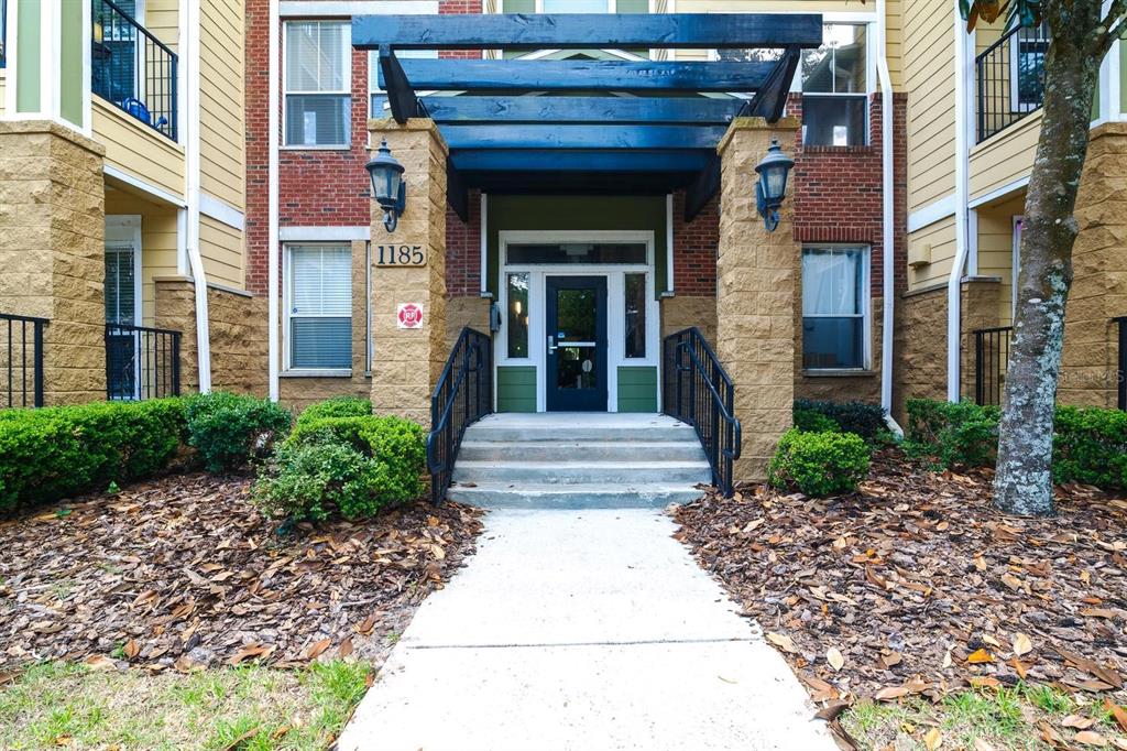 A concrete walkway leads to the entrance of a multi-story apartment building with stone pillars — 1185 SW 9TH ROAD UNIT 212, GAINESVILLE, FL