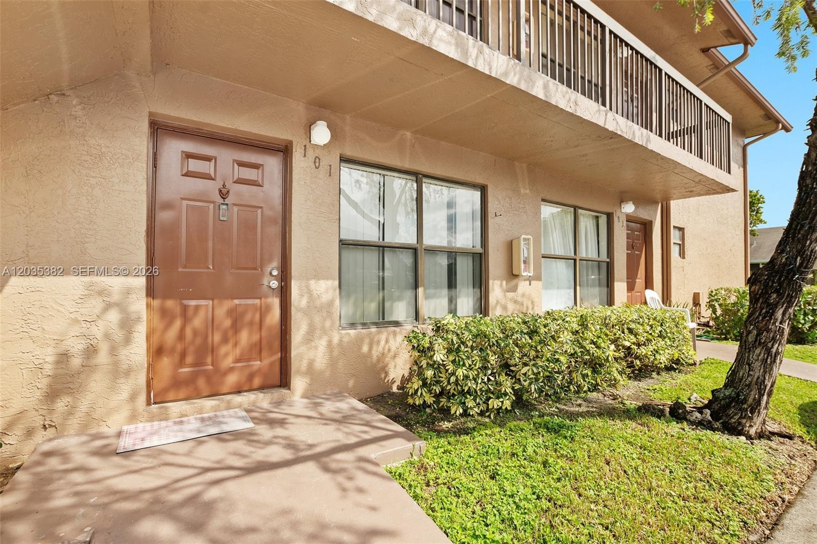 The exterior of a tan stucco apartment building features a brown front door and balcony — 10053 Winding Lake Rd # 101, Sunrise FL 33351, SUNRISE, FL