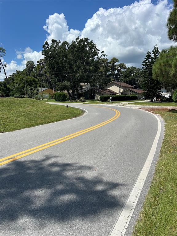 A paved road with double yellow lines curves past houses and trees under a cloudy sky — 12629 LACEY DRIVE, NEW PORT RICHEY, FL