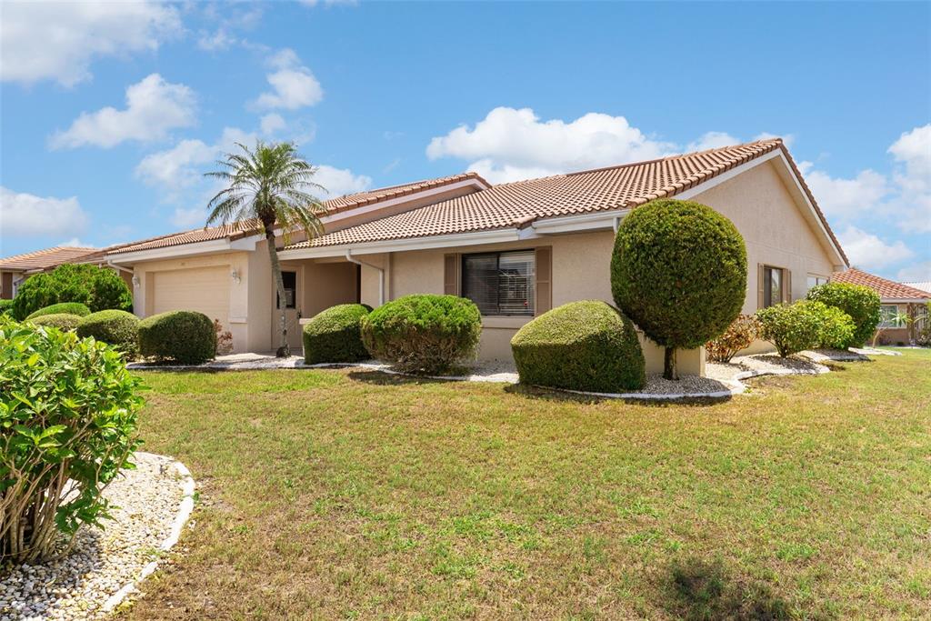 A single-story house with a tan stucco exterior, tile roof, and manicured landscaping — 701 BALTUSROL WAY, SUN CITY CENTER, FL