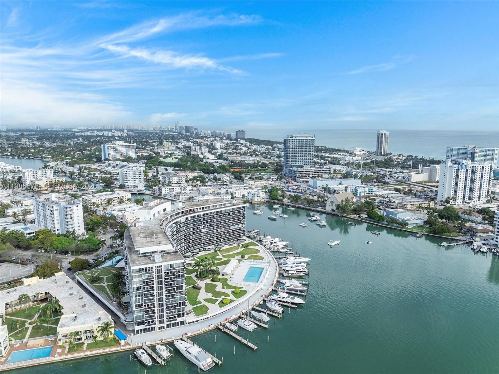 An aerial view shows a curved waterfront apartment building with a pool and boat slips — 900 Bay Dr # 411, Miami Beach FL 33141, MIAMI BEACH, FL