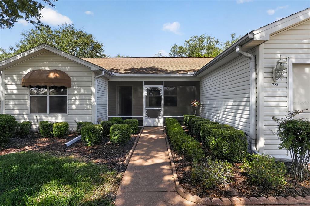 A beige-sided house features a screened front entrance, a concrete walkway, and manicured landscaping — 728 RIVERA ROAD, THE VILLAGES, FL