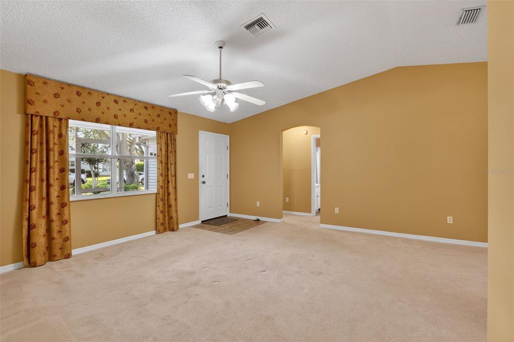 A living room with beige carpet, tan walls, a ceiling fan, and a window with curtains — 728 RIVERA ROAD, THE VILLAGES, FL