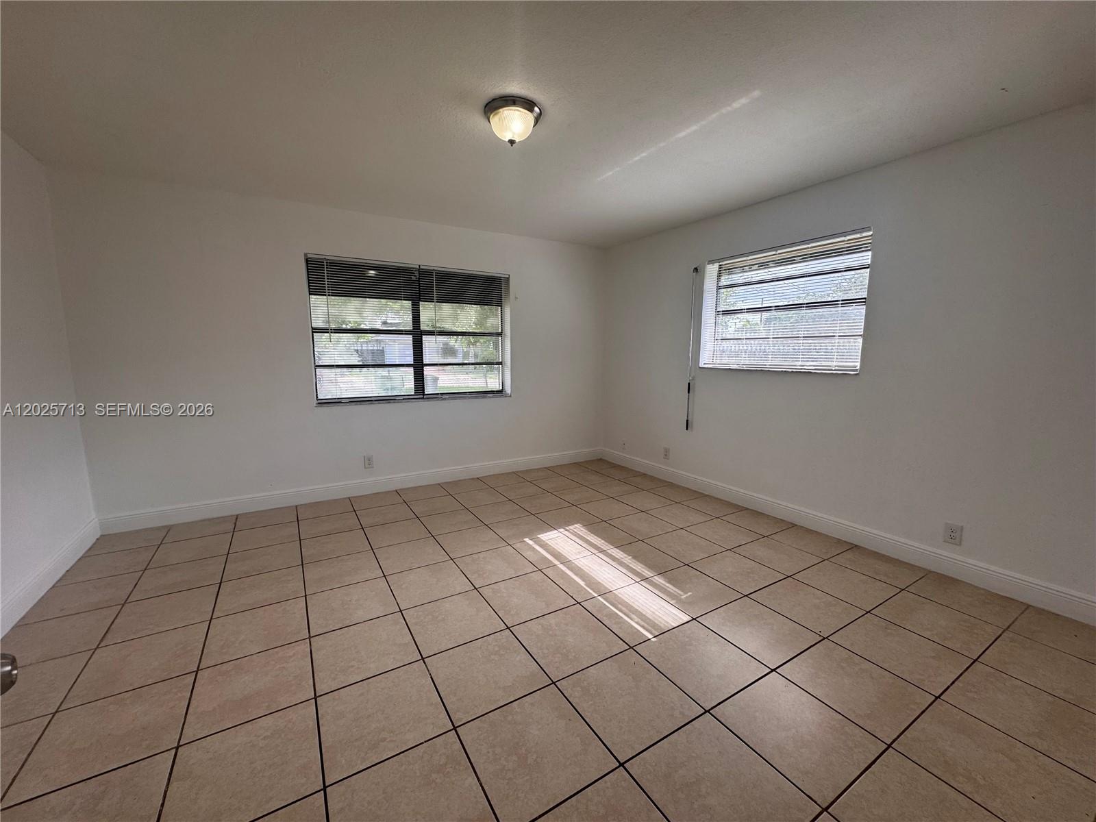 An empty room features white walls, beige tile flooring, two windows with blinds, and a ceiling light — 407 NW 7th Ct, Hallandale Beach FL 33009, HALLANDALE BEACH, FL