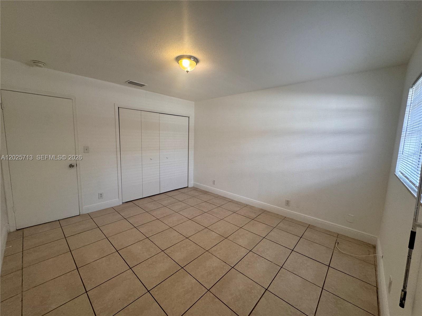 An empty room features white walls, beige tile flooring, a closet, and a window — 407 NW 7th Ct, Hallandale Beach FL 33009, HALLANDALE BEACH, FL