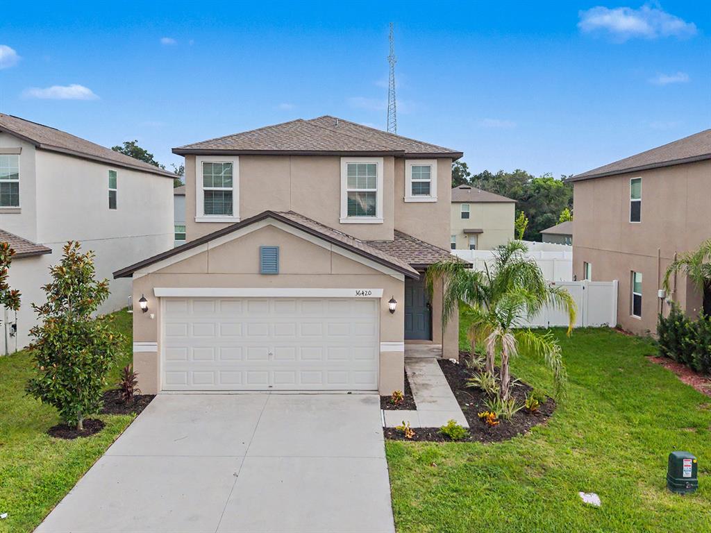 A two-story suburban house features a tan stucco exterior, a two-car garage, and landscaping — 36420 GARDEN WALL WAY, ZEPHYRHILLS, FL