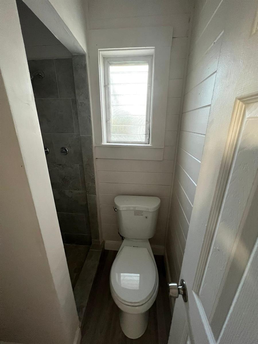 A small bathroom features a toilet, a window with white shiplap walls, and a tiled shower — 2843 Forest Hill Boulevard # 7, West Palm Beach FL 33406, WEST PALM BEACH, FL