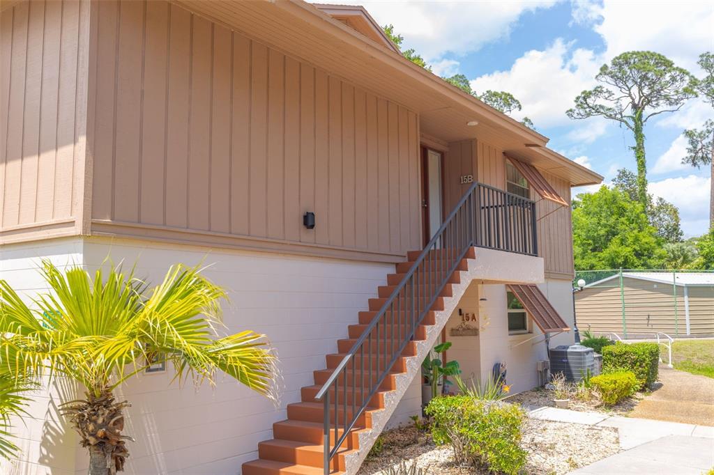 A two-story building exterior features a staircase leading to an upper-level entrance marked 15B — 180 MAGNOLIA WOODS COURT UNIT 15B, DELTONA, FL