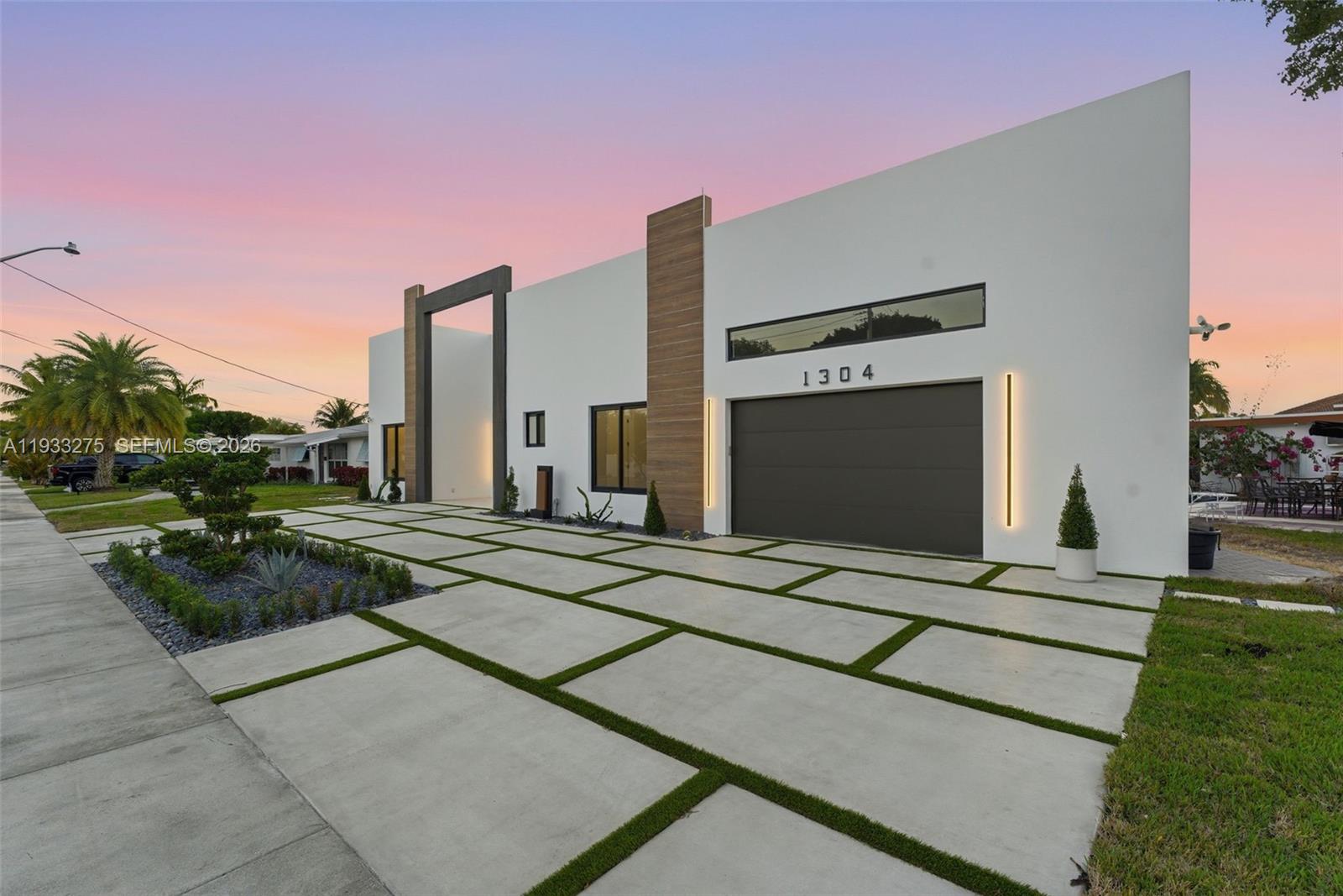 A modern white house features a large garage, wood accents, and a concrete paver driveway — 1304 SE 10th Ter, Deerfield Beach FL 33441, DEERFIELD BEACH, FL