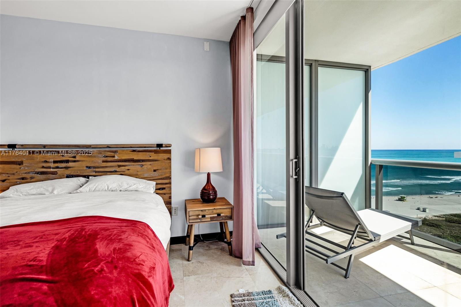 A bedroom features a rustic wooden headboard, a red blanket, and a balcony overlooking the ocean — 5875 Collins Ave # 1704, Miami Beach FL 33140, MIAMI BEACH, FL