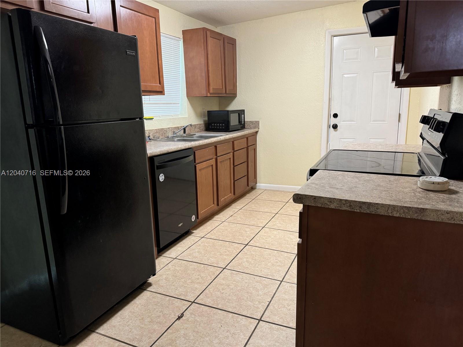 A kitchen features wood cabinets, black appliances, tan tile flooring, and a white exterior door — 9410 Gilchrist ct, Jacksonville FL 32219, JACKSONVILLE, FL