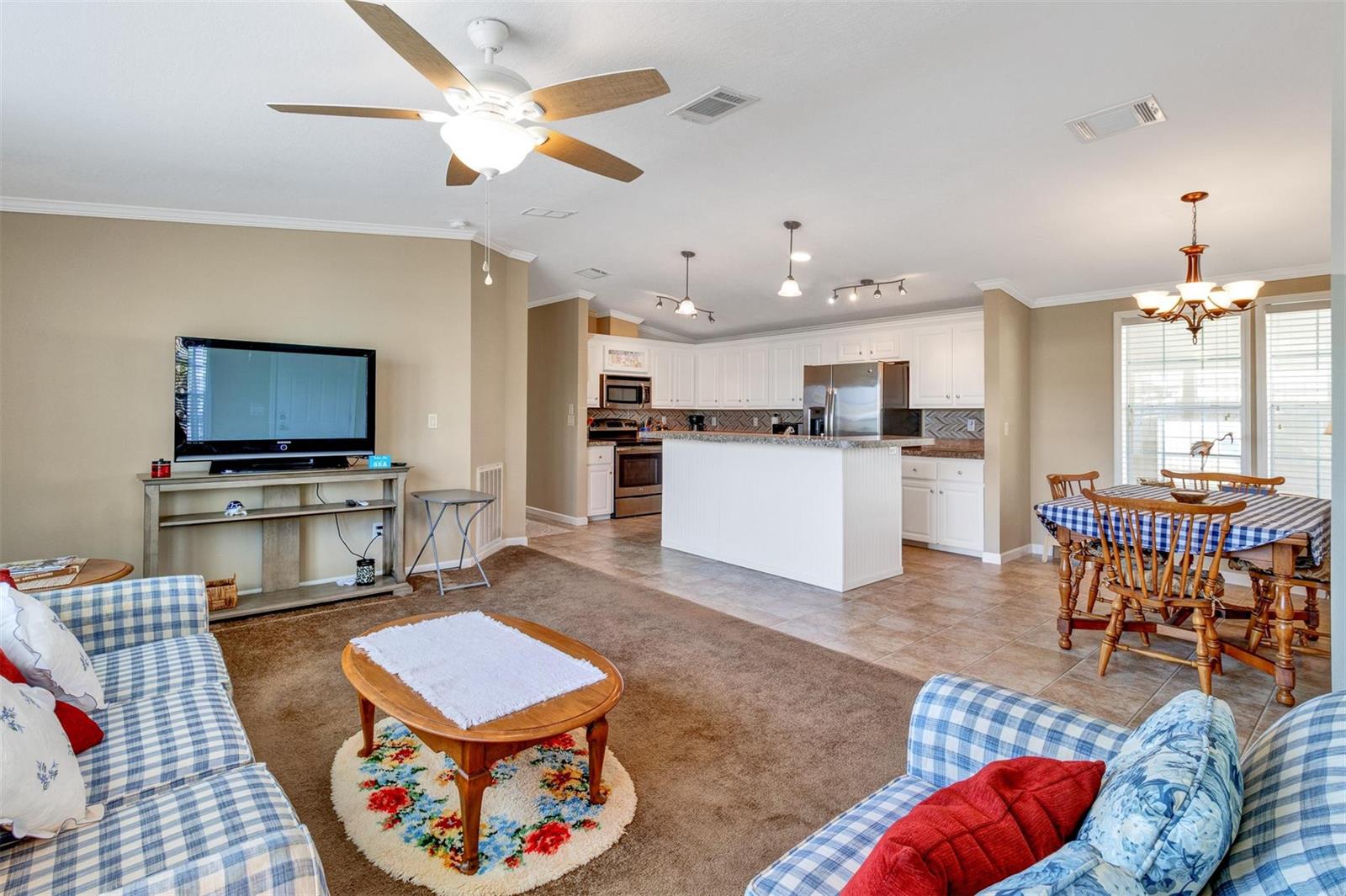 An open-concept living room and kitchen featuring a ceiling fan, brown carpet, and white cabinetry — 192 N Arbor Lane, Vero Beach FL 32960, VERO BEACH, FL