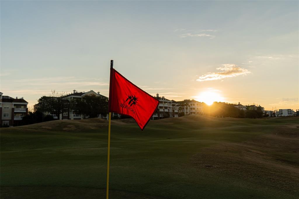 A red golf flag stands on a putting green with buildings in the background at sunset — 7636 HERITAGE CROSSING WAY, REUNION, FL