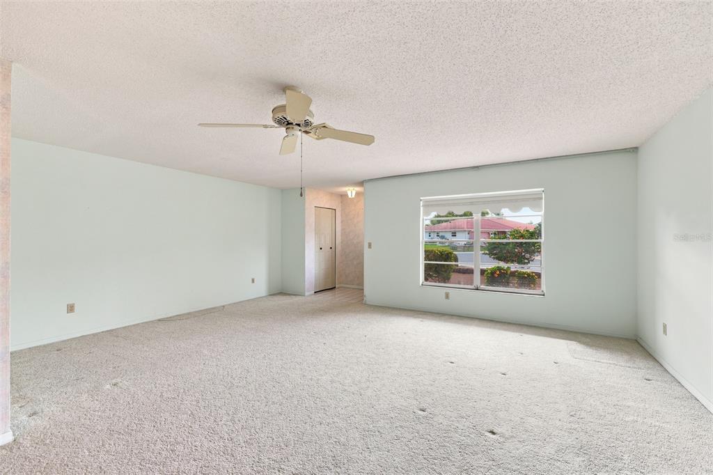 A carpeted living room features a ceiling fan, light-colored walls, and a large window — 127 WINTERSONG LANE, SUN CITY CENTER, FL