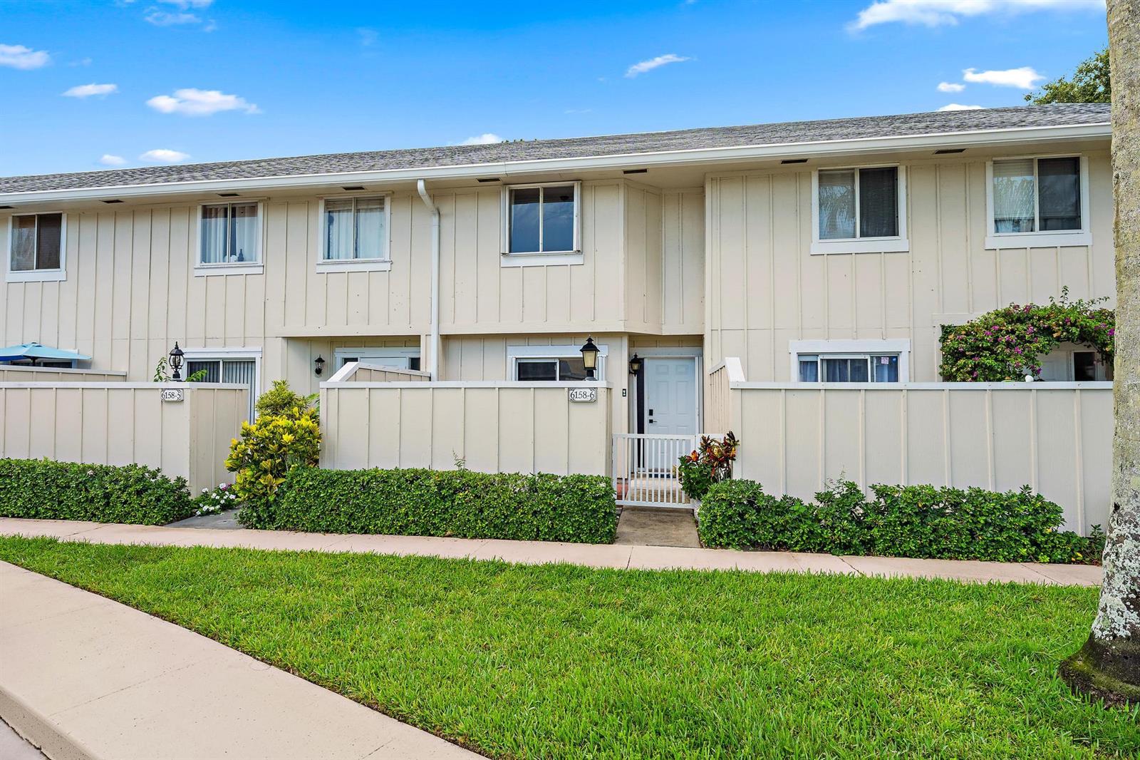 A two-story townhouse exterior features beige vertical siding, private fenced courtyards, and manicured hedges — 6158 Riverwalk Lane # 6, Jupiter FL 33458, JUPITER, FL