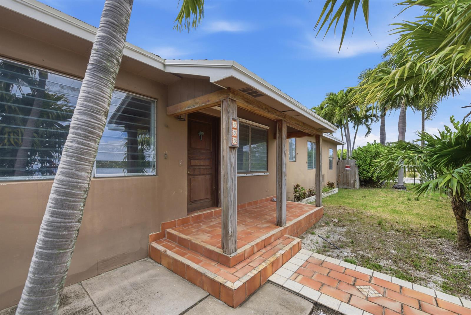 A tan house exterior features a tiled front porch with wooden posts and palm trees — 303 Cornell Drive, FL 33460, FL