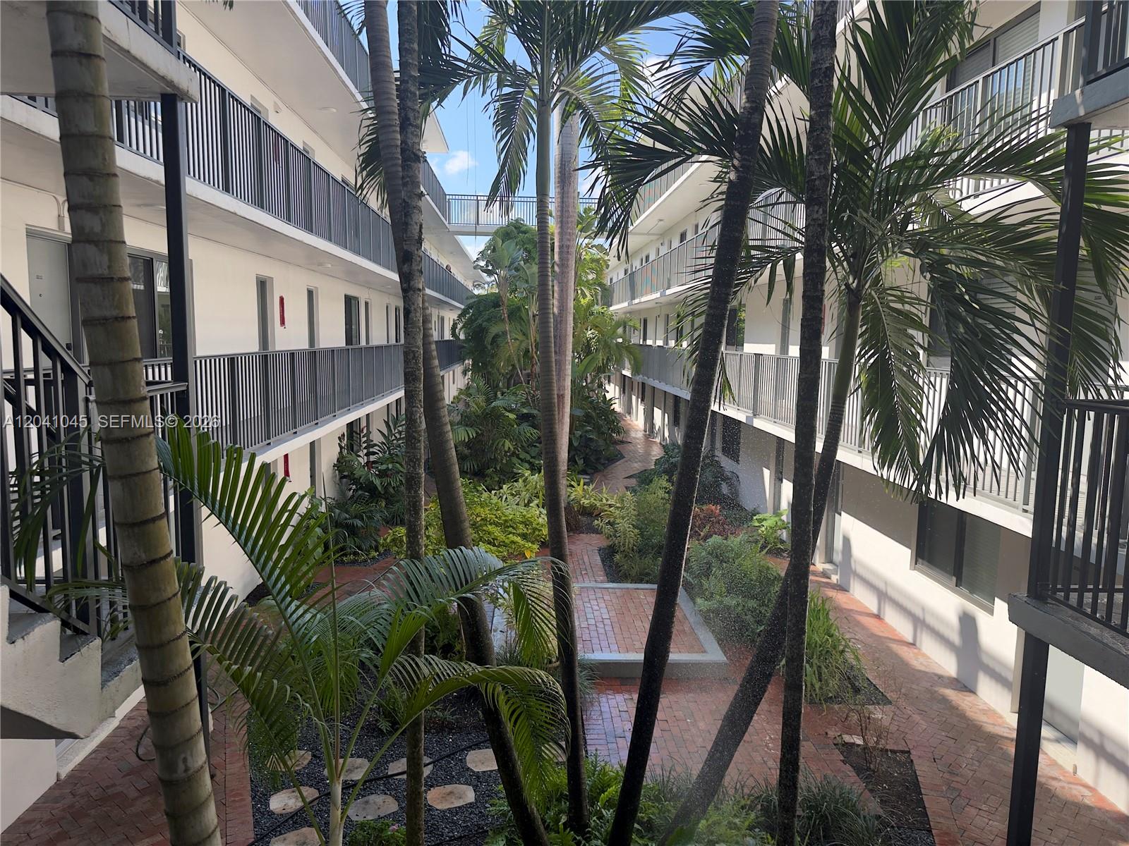 A courtyard with brick walkways and palm trees is surrounded by a multi-story apartment building — 3242 Mary St # S115, Miami FL 33133, MIAMI, FL