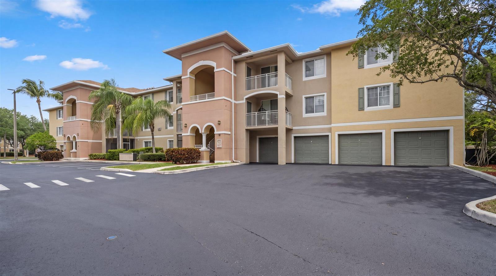 The exterior of a multi-story apartment building features tan stucco walls and attached ground-level garages — 6475 Emerald Dunes Drive # 206, West Palm Beach FL 33411, WEST PALM BEACH, FL