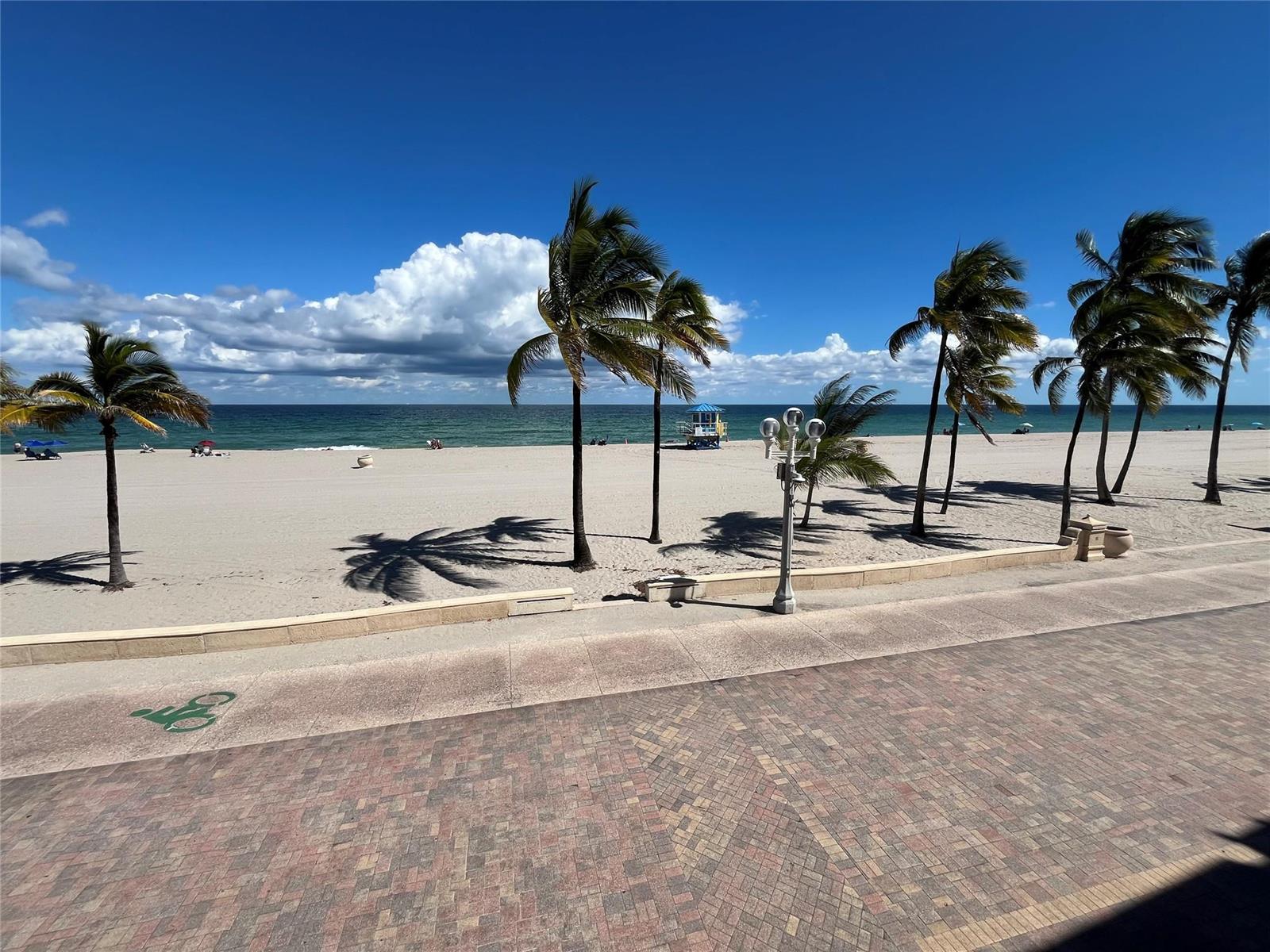 A view of a sandy beach with palm trees, a lifeguard stand, and a brick walkway — 310 S Broadwalk # 3b, Hollywood FL 33019, HOLLYWOOD, FL