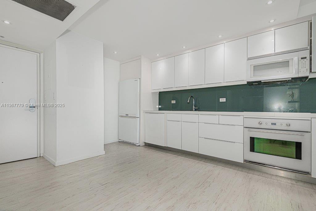 A modern kitchen features white flat-panel cabinetry, a green glass backsplash, and light wood flooring — 2301 Collins Ave # 1032, Miami Beach FL 33139, MIAMI BEACH, FL