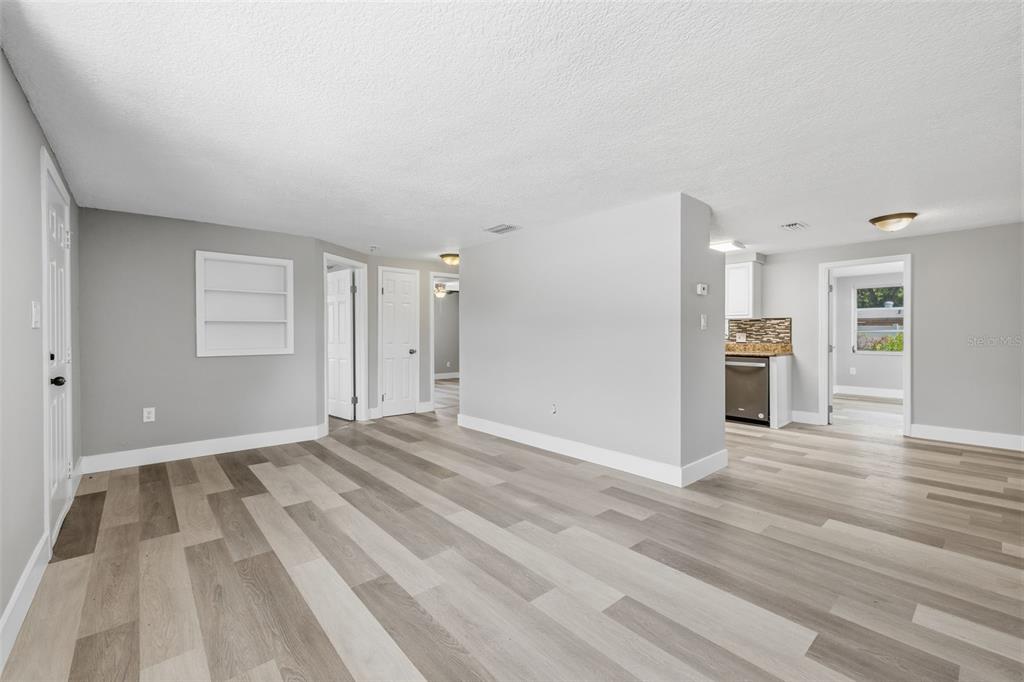 An open-concept living area features light gray walls, wood-look flooring, and a kitchen beyond — 6150 EMERALD DRIVE, NEW PORT RICHEY, FL