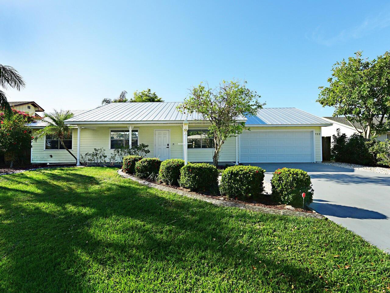 A single-story house with light yellow siding, a metal roof, and a paved driveway — 598 NE Maranta Terrado, Jensen Beach FL 34957, JENSEN BEACH, FL