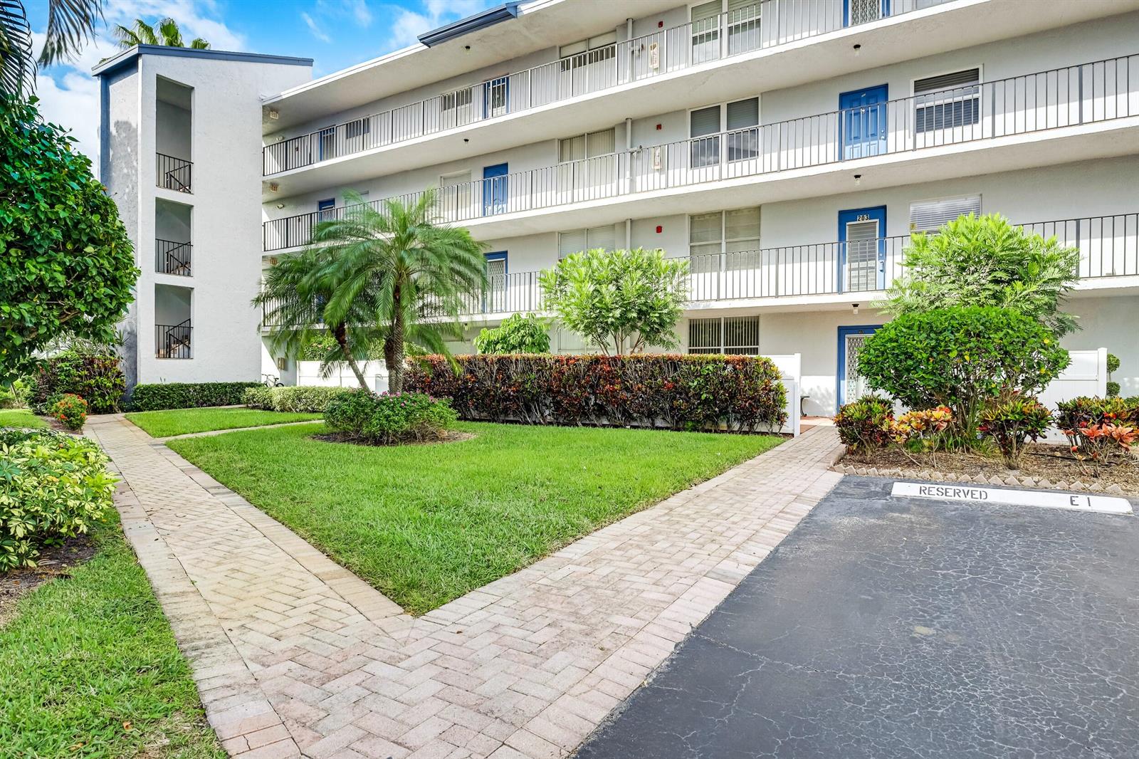 A multi-story apartment building features white exterior walls, blue doors, and a landscaped courtyard — 6650 S Oriole Boulevard # 1030, Delray Beach FL 33446, DELRAY BEACH, FL