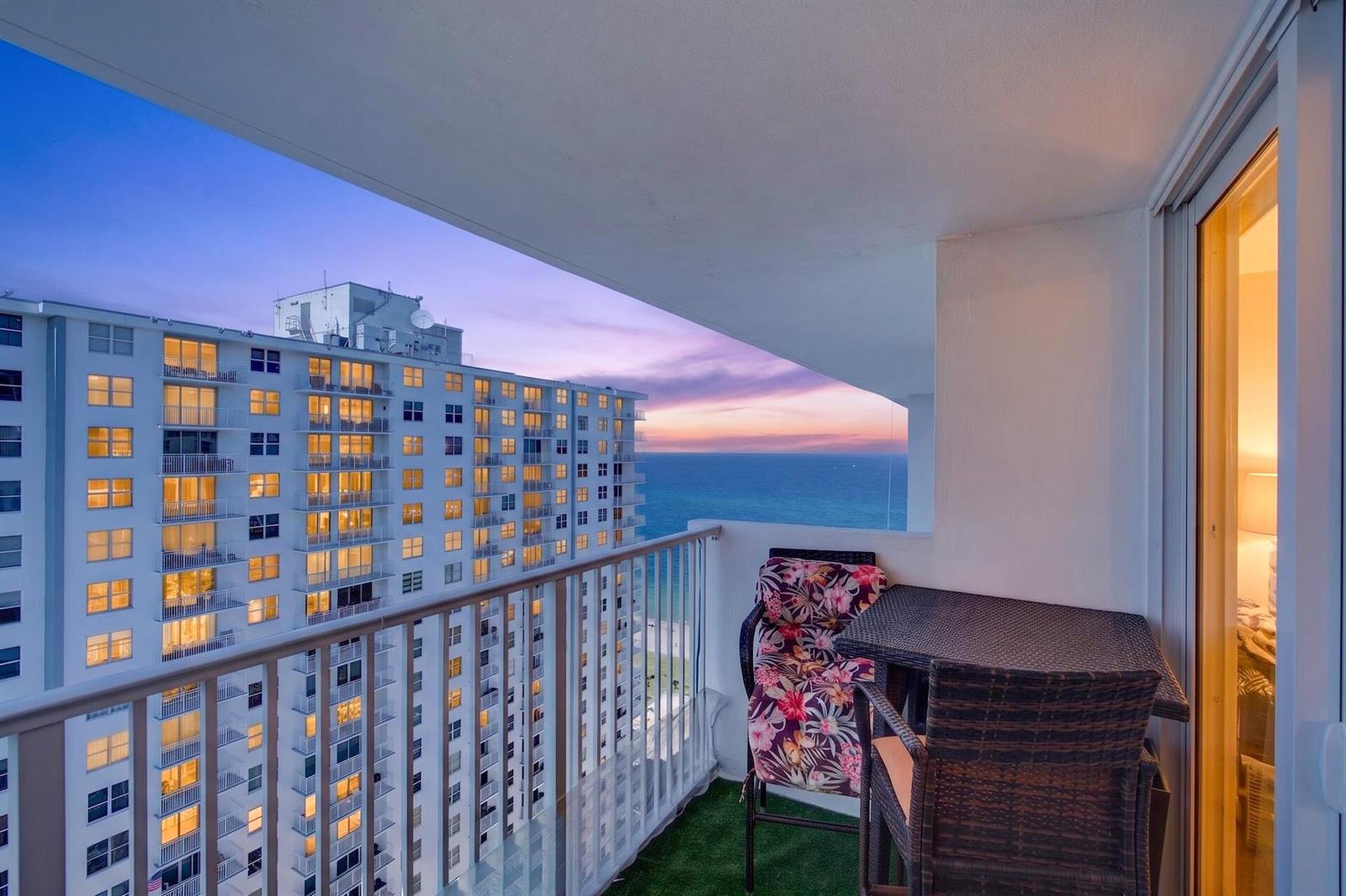 A balcony with wicker furniture overlooks a neighboring high-rise building and the ocean at sunset — 111 Briny Avenue # 2711, Pompano Beach FL 33062, POMPANO BEACH, FL