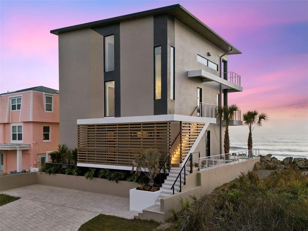 A modern three-story house with a slatted wood privacy screen and exterior staircase at sunset — 6887 S ATLANTIC AVENUE, NEW SMYRNA BEACH, FL