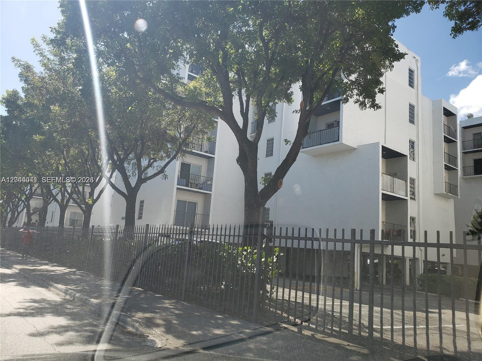 A multi-story white apartment building stands behind a metal fence and large trees — 4550 W 16th Ave # 301, Hialeah FL 33012, HIALEAH, FL