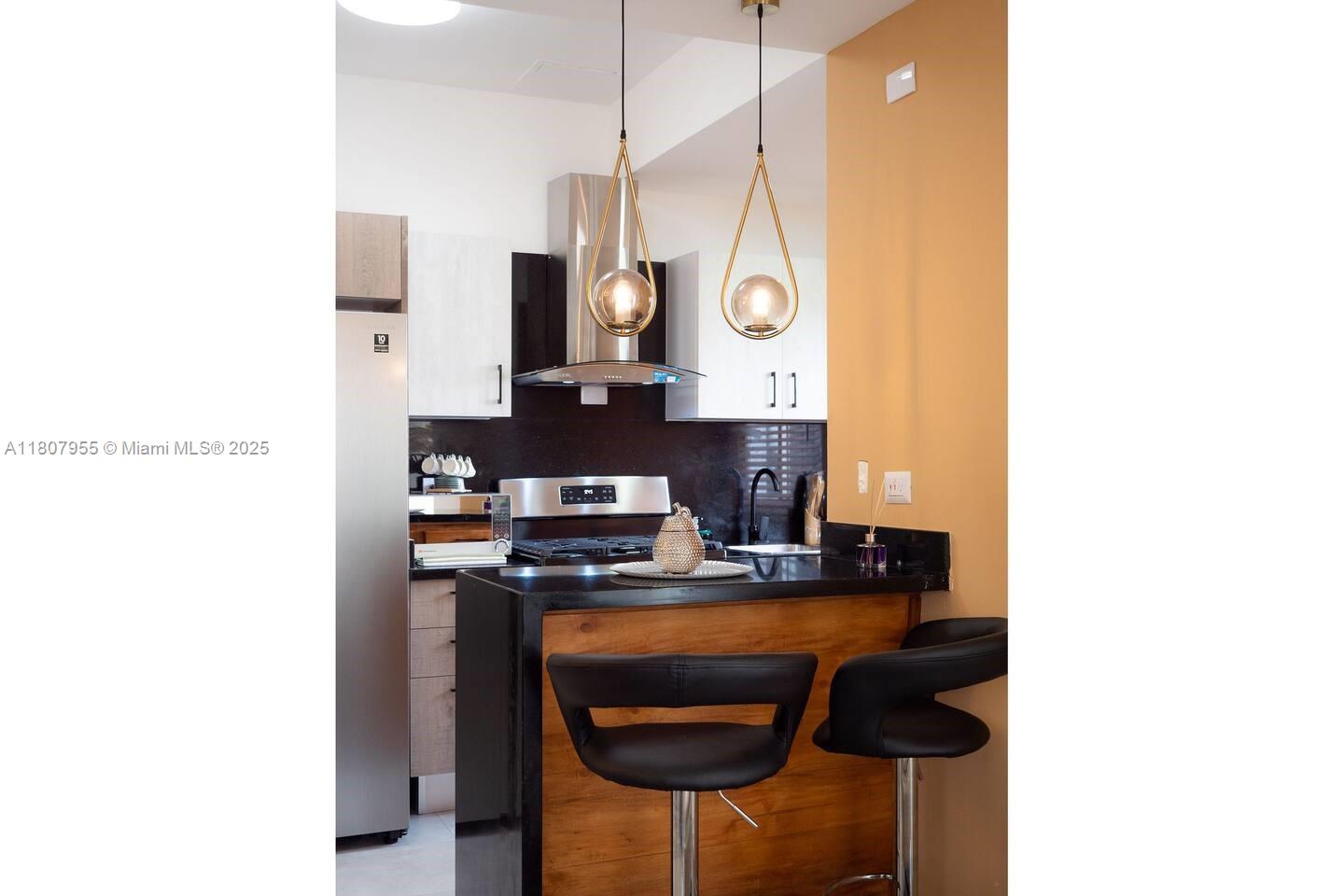A kitchen features a black countertop, wooden breakfast bar, two pendant lights, and stainless appliances — Julio Aibar #39 Suites By Refa Piantini # 401, RD, RD