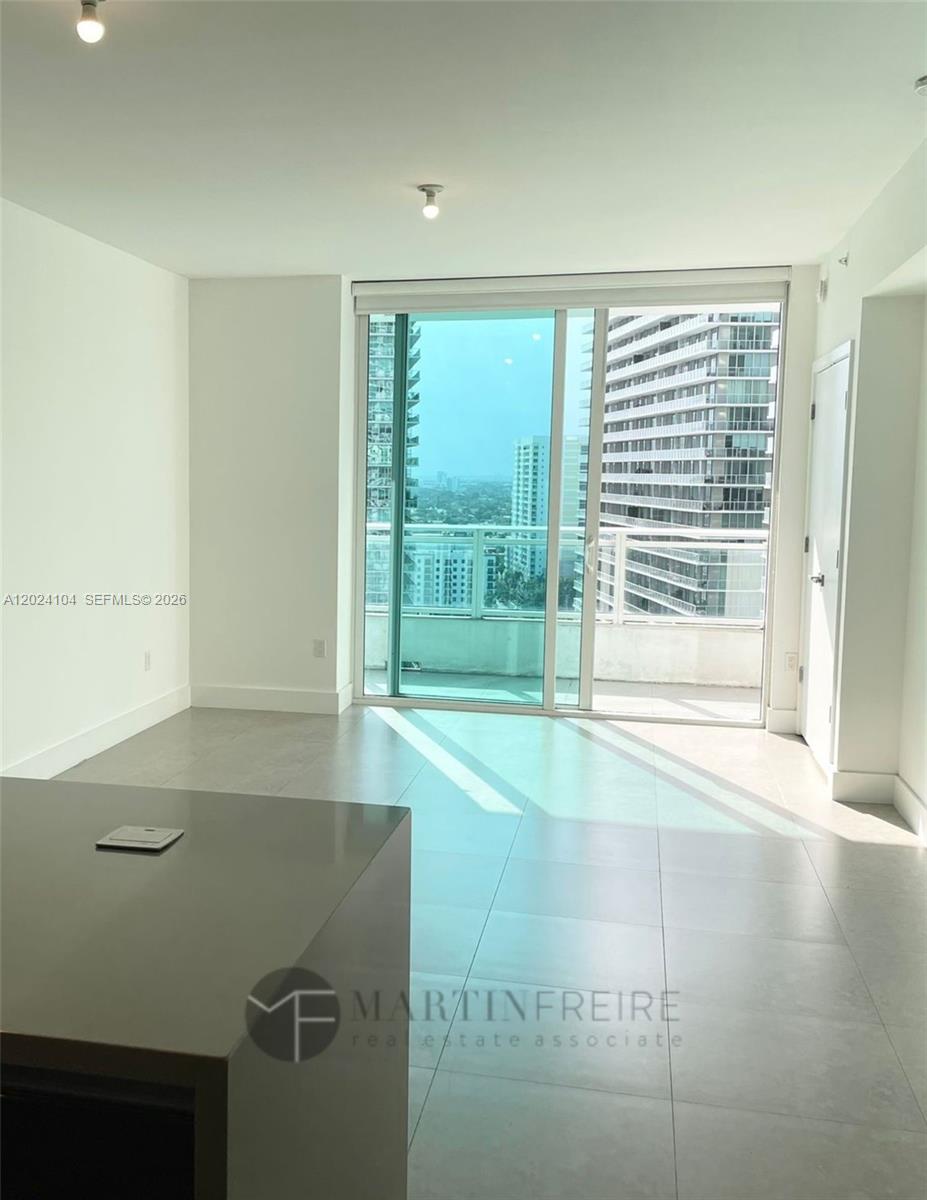 A bright, unfurnished room features light tile flooring, a kitchen island, and a glass balcony door — 1080 Brickell Ave # 2005, Miami FL 33131, MIAMI, FL