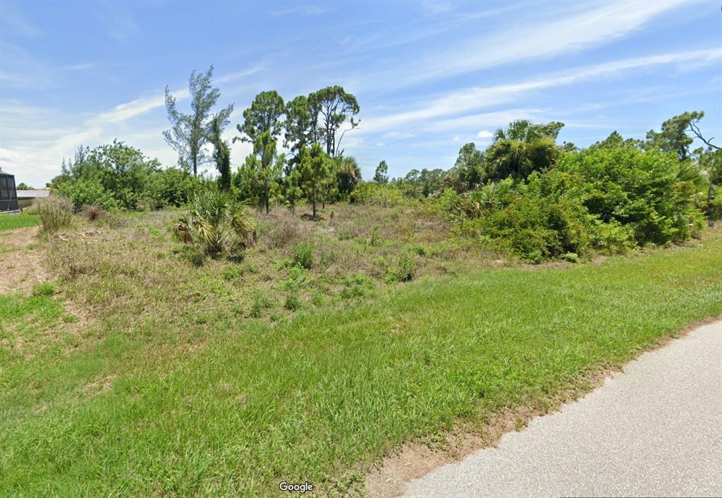 A vacant, grassy lot with scattered trees and shrubs sits beside a paved road — 15664 ALDAMA CIRCLE, PORT CHARLOTTE, FL