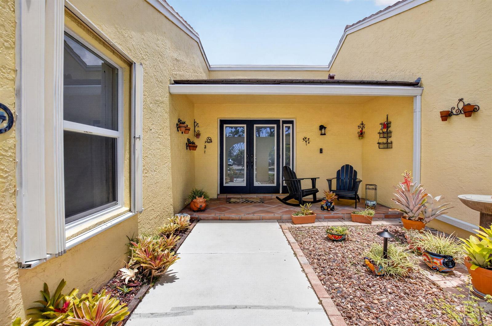 A yellow stucco house features a front courtyard with a concrete walkway and potted plants — 2305 SW 23rd Cranbrook Drive, Boynton Beach FL 33436, BOYNTON BEACH, FL