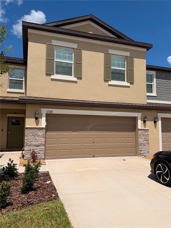 A two-story townhouse exterior features a tan stucco facade, a two-car garage, and stone accents — 17747 TAWNY MALT PLACE, LAND O LAKES, FL