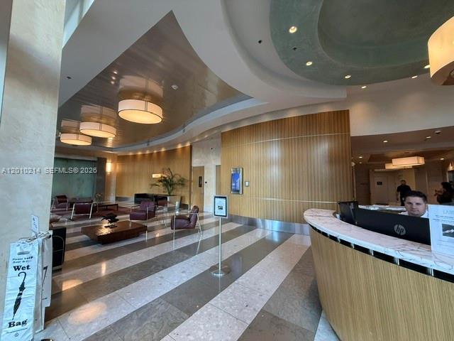 A modern hotel lobby features a curved reception desk, wood-paneled walls, and recessed ceiling lighting — 175 SW 7th St # 1605, Miami FL 33130, MIAMI, FL