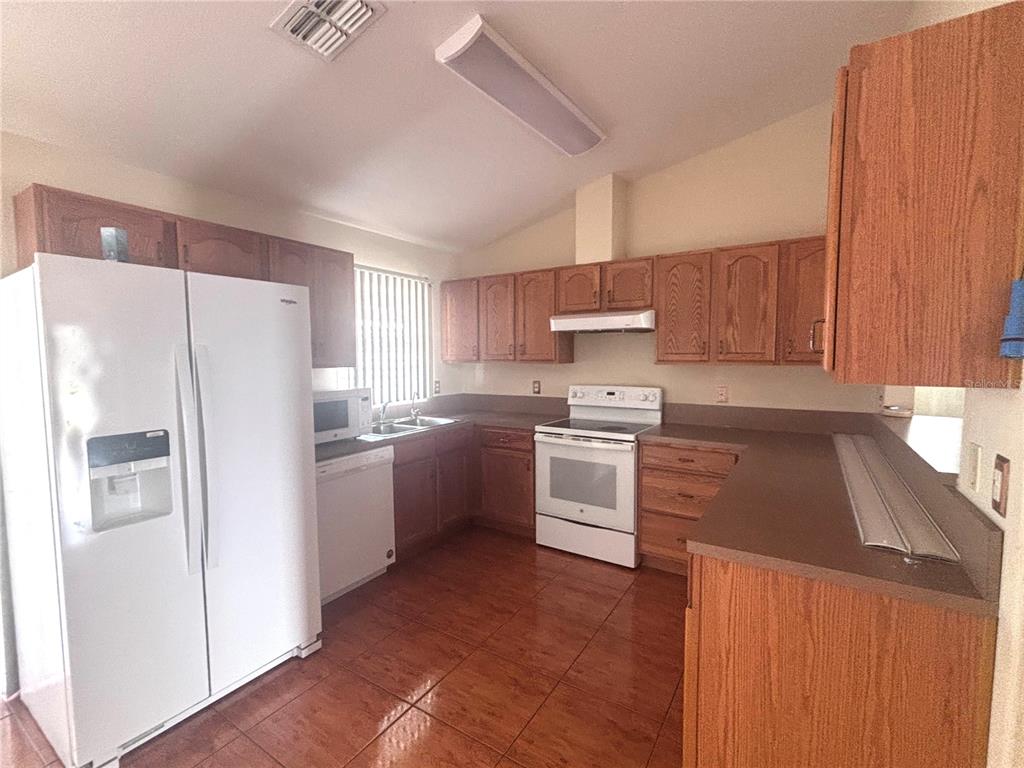 A kitchen features wood cabinets, white appliances, brown countertops, and brown tile flooring — 1912 MANATEE DR, POINCIANA, FL