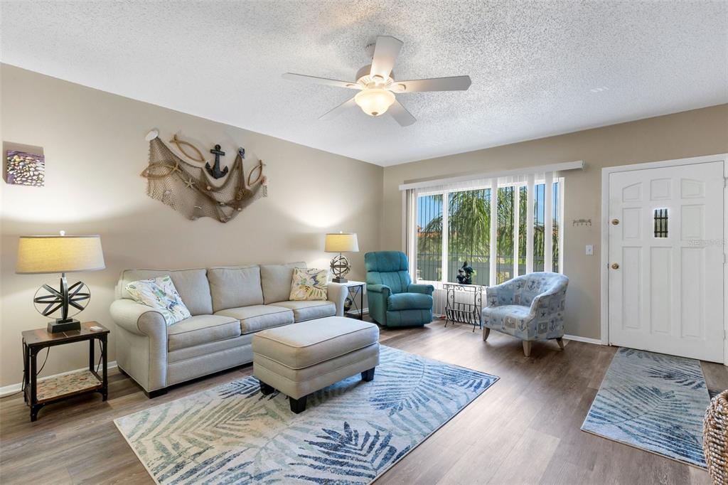 A living room features a beige sofa, matching ottoman, blue armchair, and coastal-themed wall decor — 2401 ECUADORIAN WAY UNIT 57, CLEARWATER, FL