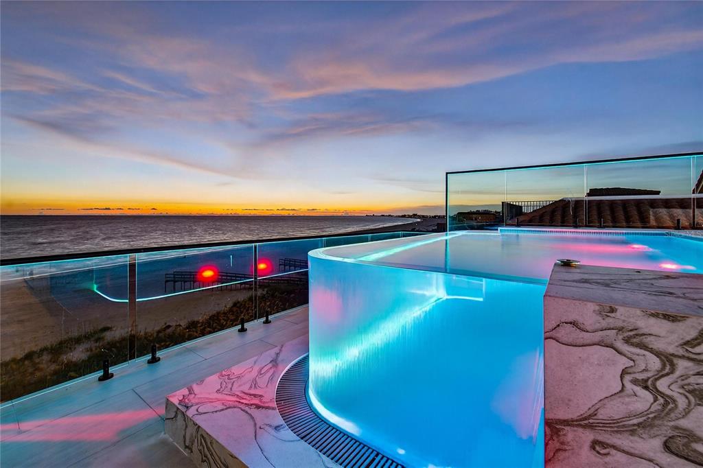 An illuminated rooftop infinity pool overlooks the ocean at sunset with a glass balcony railing — 9600 W GULF BOULEVARD, TREASURE ISLAND, FL