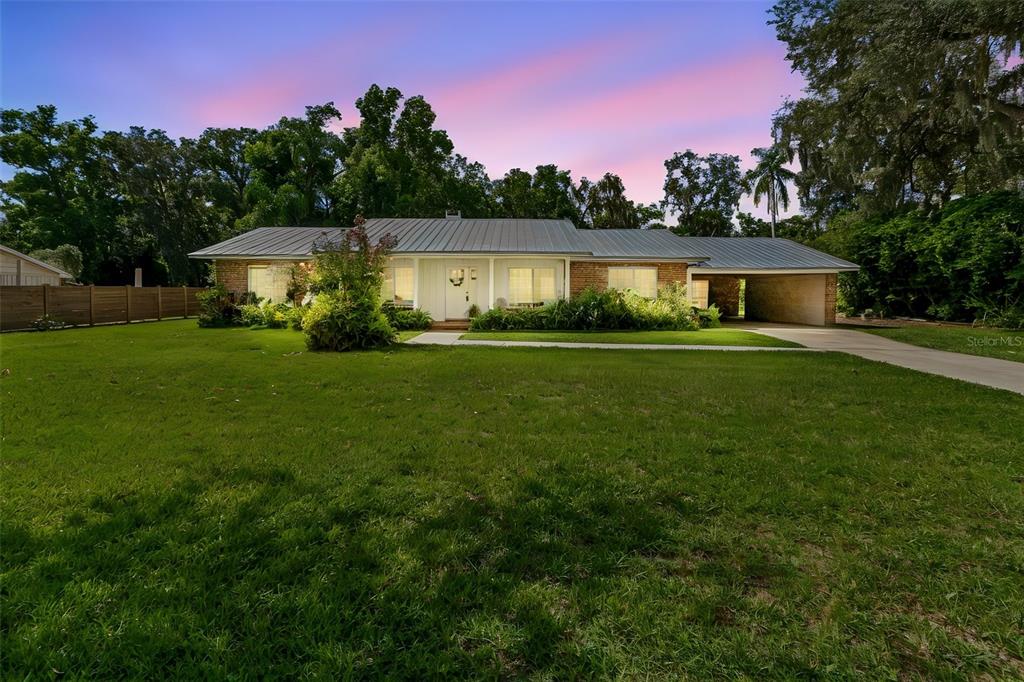 A single-story brick house with a metal roof and a carport sits on a lawn — 909 TOMLINSON DRIVE, LUTZ, FL