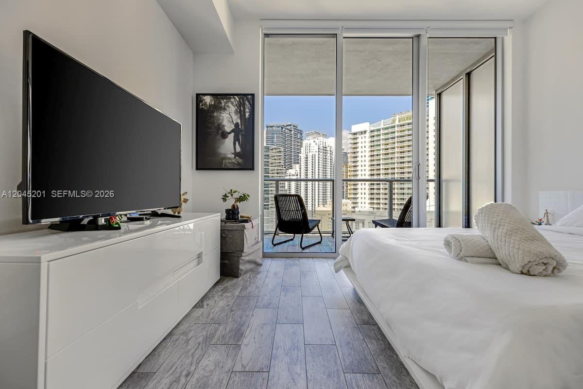A bedroom with a large television, white dresser, and balcony access overlooking city buildings — 1300 Brickell Bay Dr # 2713, Miami FL 33131, MIAMI, FL