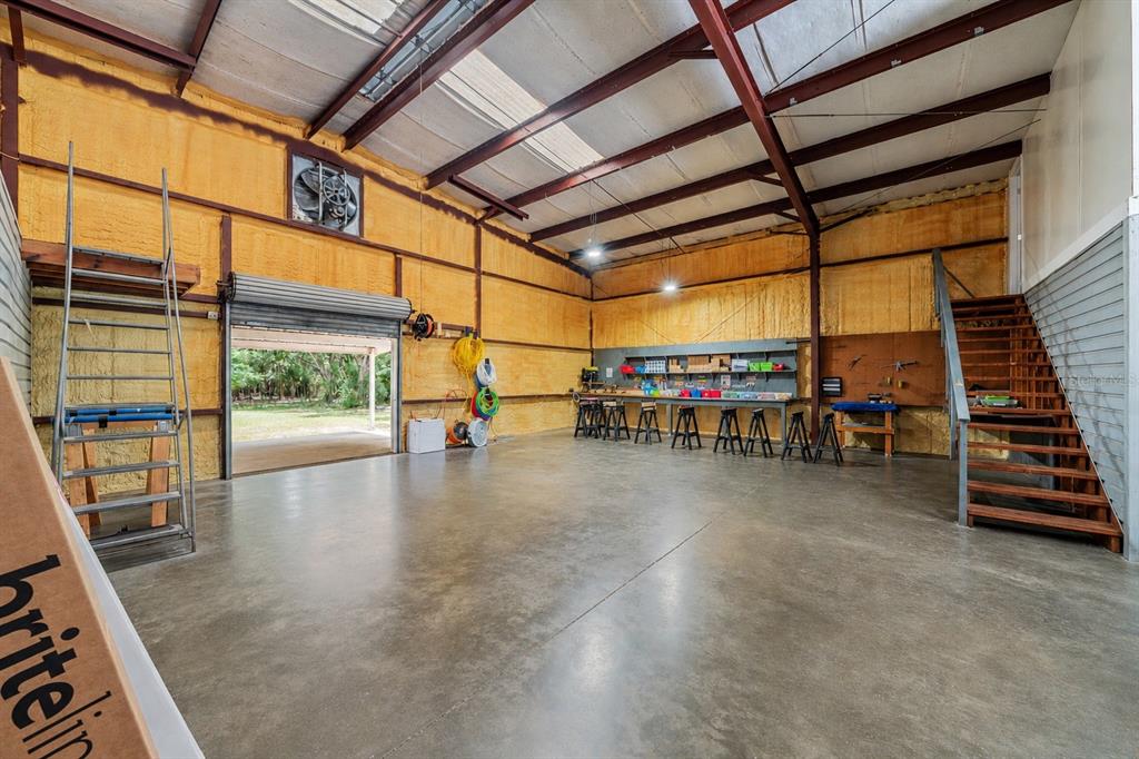 A large, open workshop with concrete floors, yellow spray foam insulation, and a wooden staircase — 15725 SE 36TH AVENUE, SUMMERFIELD, FL