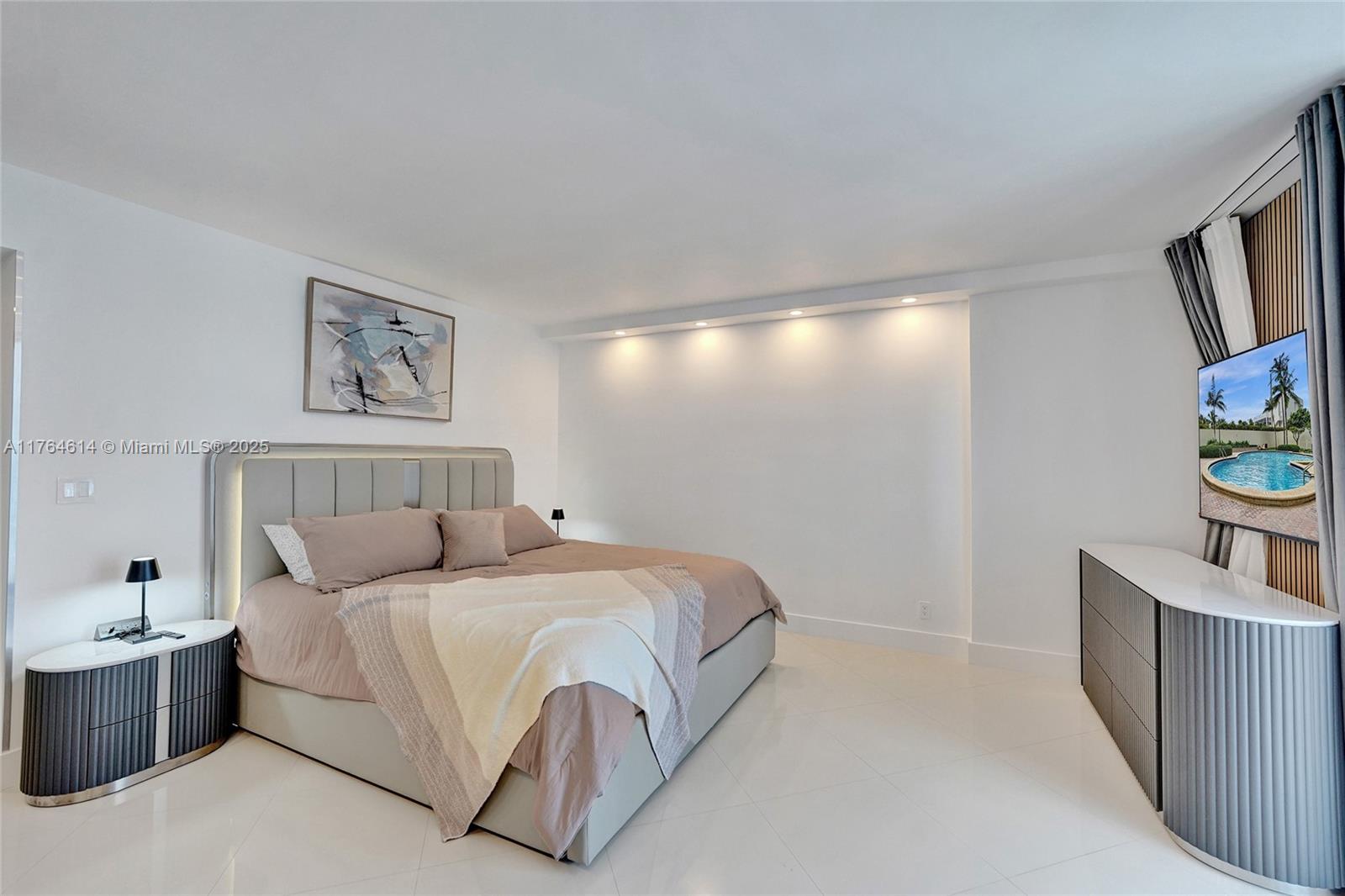 A modern bedroom features a gray upholstered bed, matching nightstands, and a wall-mounted television — 4001 S Ocean Dr # 16P, Hollywood FL 33019, HOLLYWOOD, FL