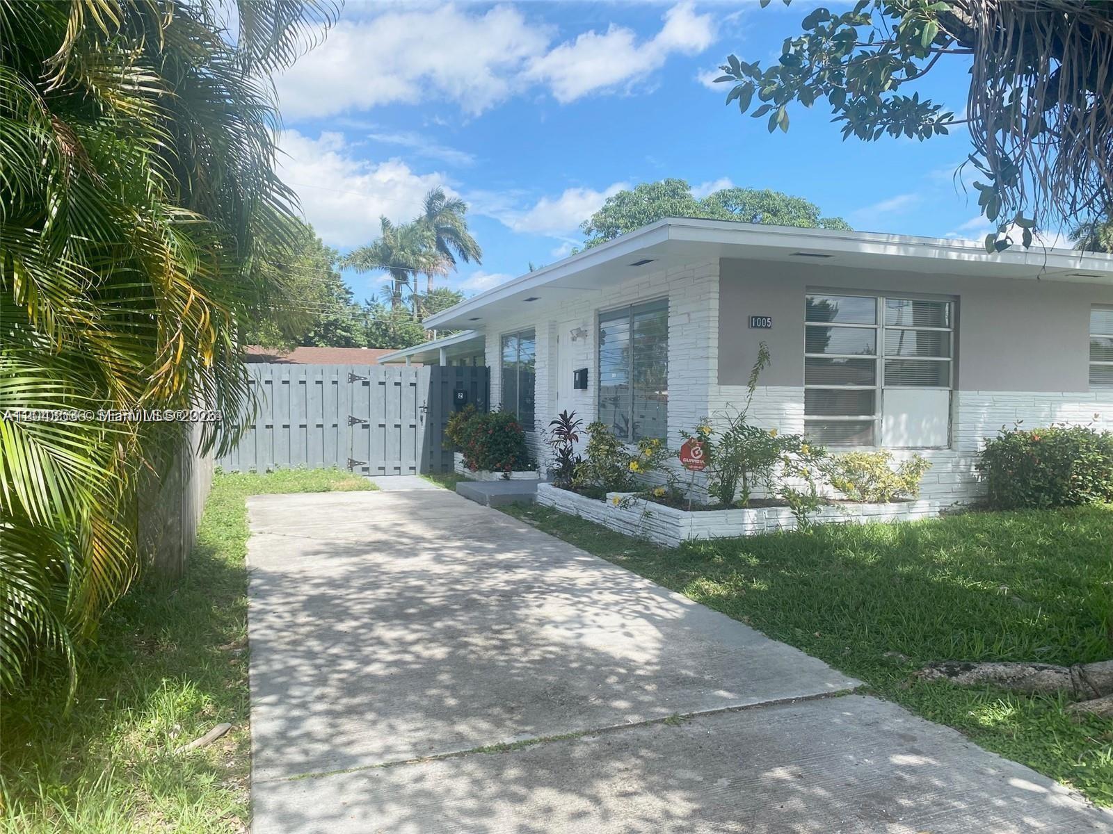 A single-story house with a white brick facade, concrete driveway, and a gray wooden fence — 1005 NE 2nd St, Hallandale Beach FL 33009, HALLANDALE BEACH, FL