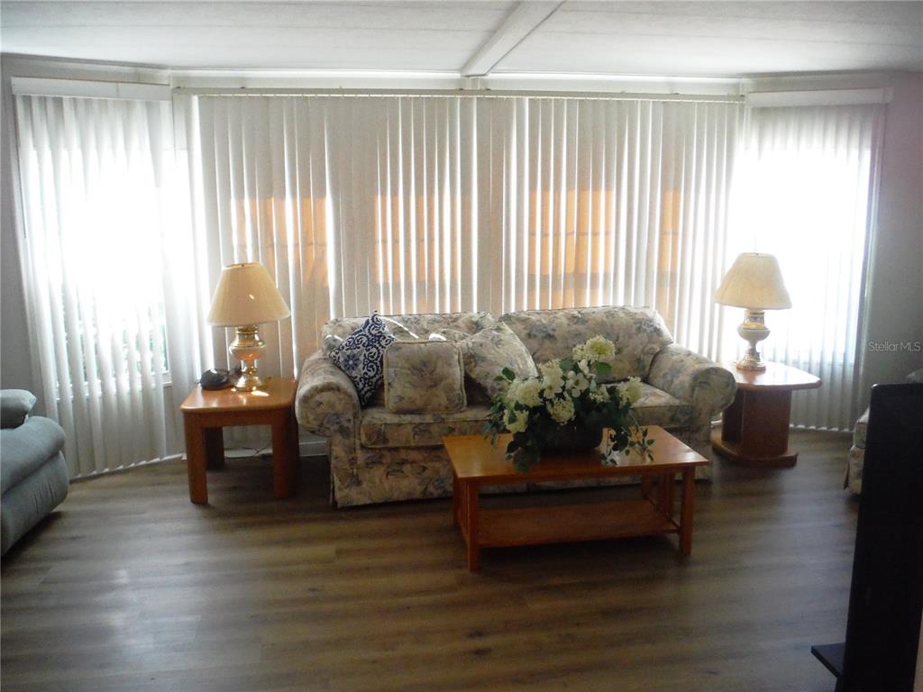 A living room features a floral sofa, wooden tables, lamps, and vertical blinds on windows — 141 FOREST LANE UNIT 13, SAFETY HARBOR, FL