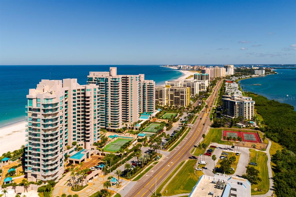 An aerial view shows high-rise beachfront condominiums, a coastal road, and tennis courts — 1560 GULF BOULEVARD UNIT 1507, CLEARWATER BEACH, FL