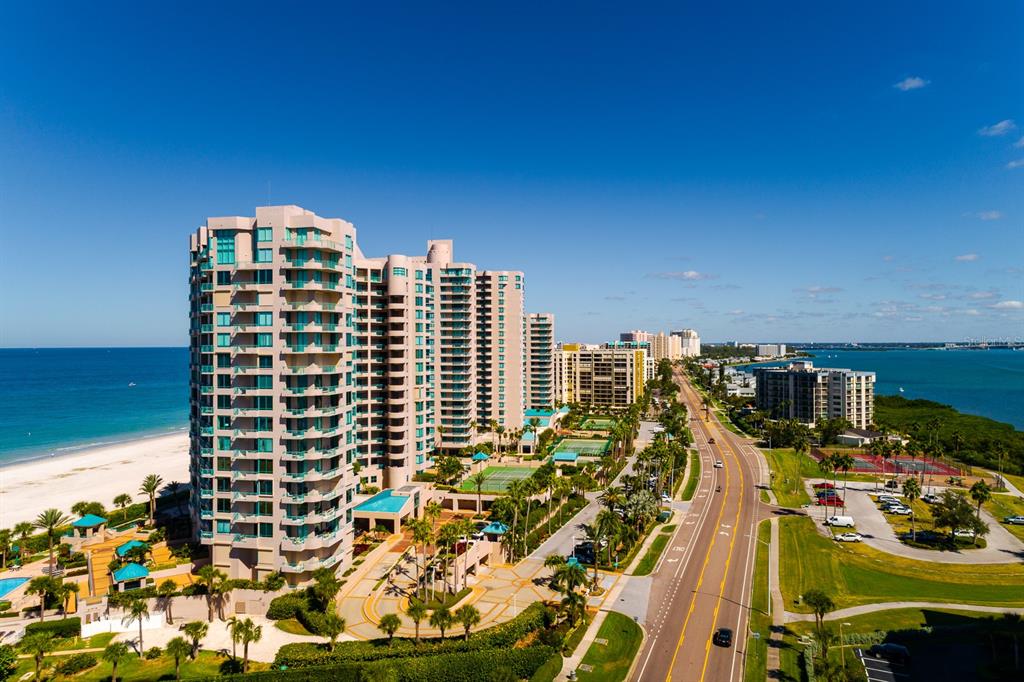An aerial view shows a tall beachfront condominium complex beside a road and the ocean — 1560 GULF BOULEVARD UNIT 1507, CLEARWATER BEACH, FL