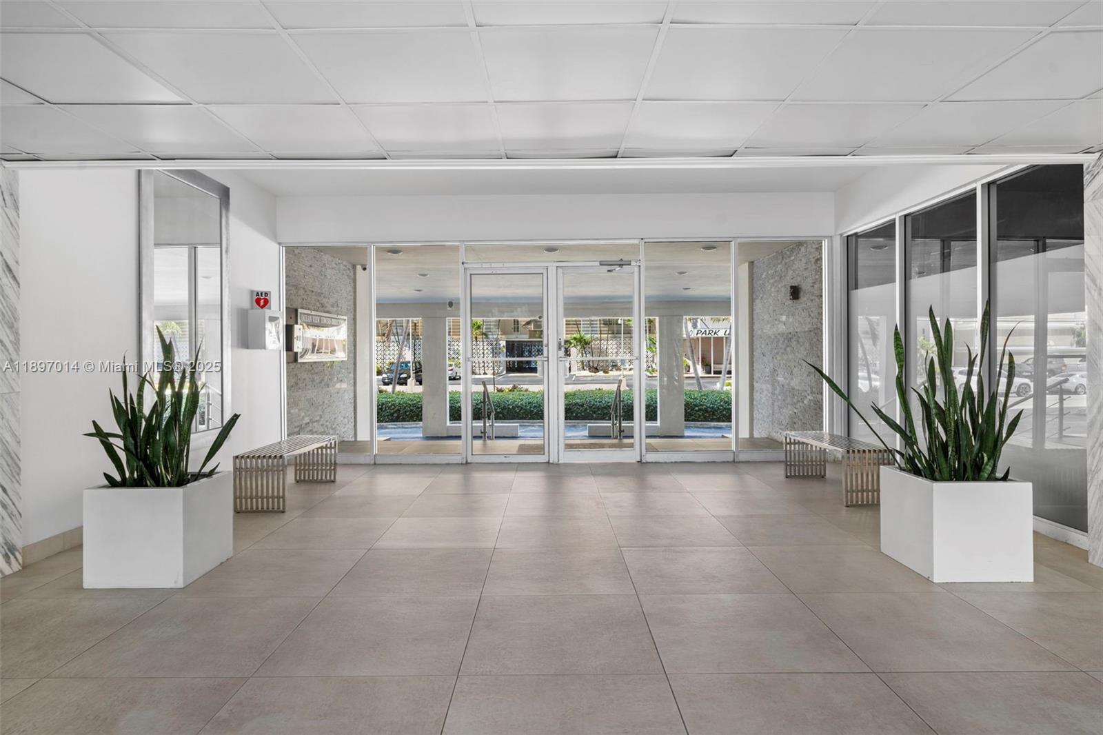 A bright building lobby features tile floors, two large potted plants, and glass entrance doors — 401 Golden Isles Dr # 304, Hallandale Beach FL 33009, HALLANDALE BEACH, FL